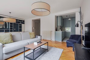 Ein modernes Wohnzimmer mit einer Couch, einem Couchtisch und einem Fernseher. Durch einen offenen Durchgang ist eine Küche sichtbar.