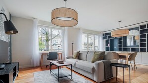 Een moderne woonkamer met een bank, salontafel en een eethoek. Er zijn grote ramen, een boekenkast en een televisie.