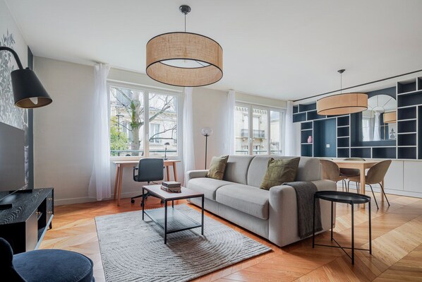 A modern living room with a sofa, coffee table, and dining area. There are large windows, a bookshelf, and a TV. - Blueground | Paris, view, nr Parc de la Villette (Paris)