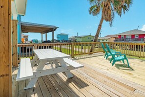 Eine Holzterrasse mit Picknicktischen und -stühlen, einem überdachten Sitzbereich und Blick auf das Meer und Gebäude im Hintergrund.