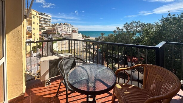 Ein Balkon mit Glastisch und Korbstühlen, der auf eine Küstenstadt mit Gebäuden und einem Strand blickt.