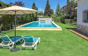Una piscina con dos sillas de descanso y una sombrilla, rodeada de césped cuidado y árboles.