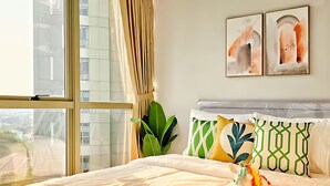 A modern hotel room with a large bed, a view of the city through floor-to-ceiling windows, and a cityscape visible outside. - Vastu By SanLiving - 2BR - HubLife Mall (Jakarta)