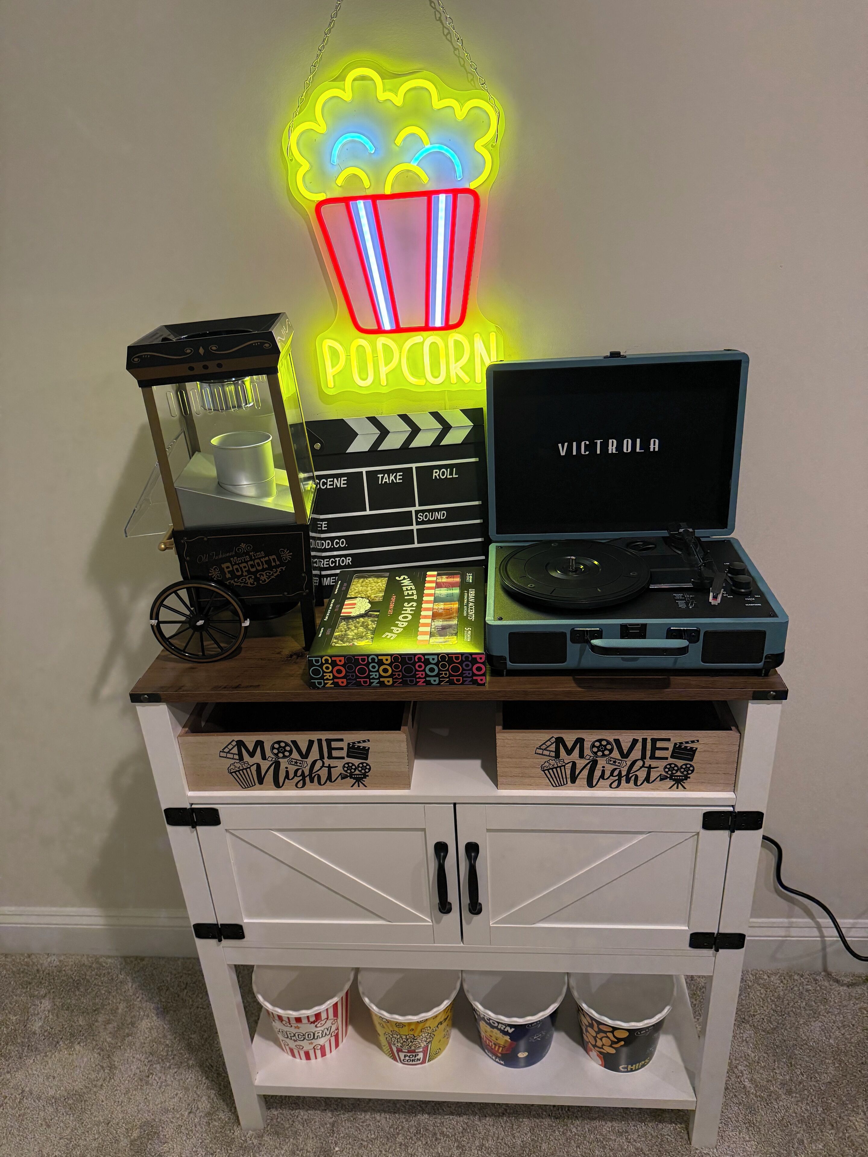 Ein Stauraum mit leuchtendem Neon-Popcorn-Schild, einem Filmklapperbrett, einem Plattenspieler und beschrifteten Schubladen für ‚Filmabend‘.
