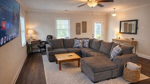 Uma sala de estar com sofá seccional, mesa de centro, ventilador de teto e uma televisão grande na parede.