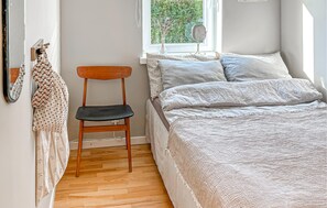 Una camera da letto con una sedia in legno, un letto con coperta trapuntata, una finestra vista verde e uno specchio appeso al muro.