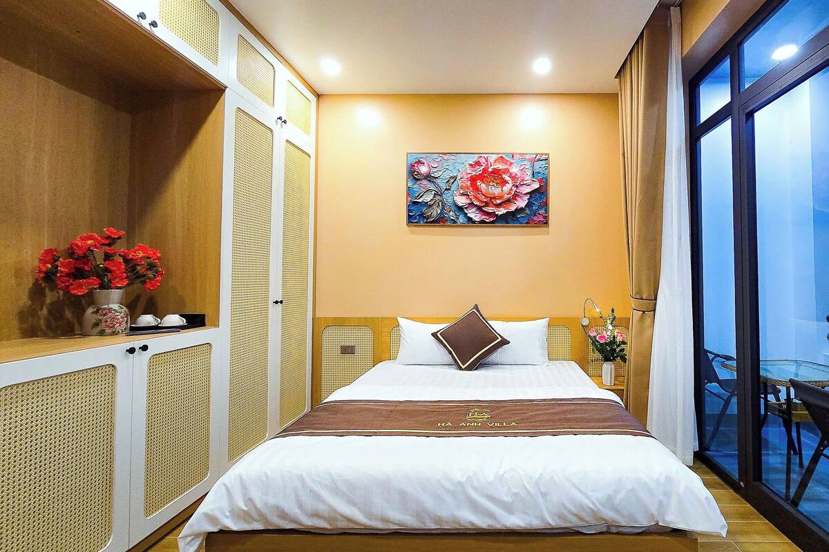 A hotel room with a bed, a painting, a vase of flowers, and a view of the outside.