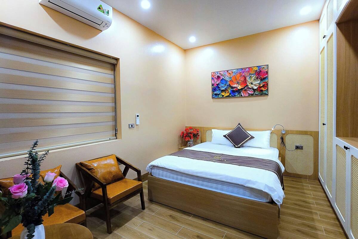 A bedroom with a bed, a chair, a window with blinds, a painting on the wall, and an air conditioning unit.