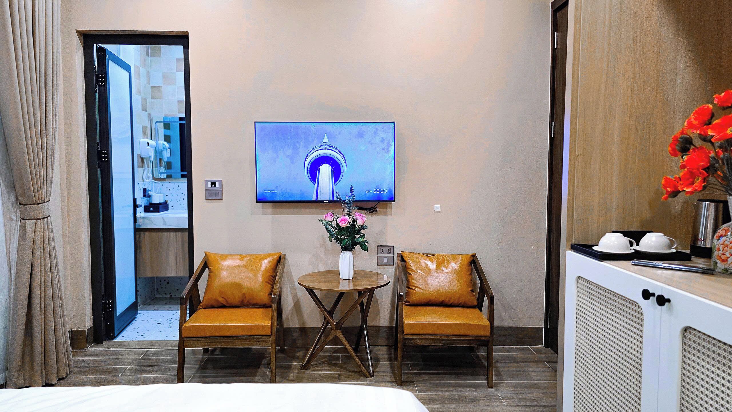 Habitación de hotel con una cama, dos sillones de cuero, una mesita con un florero, un televisor en la pared y una zona de kitchenette.
