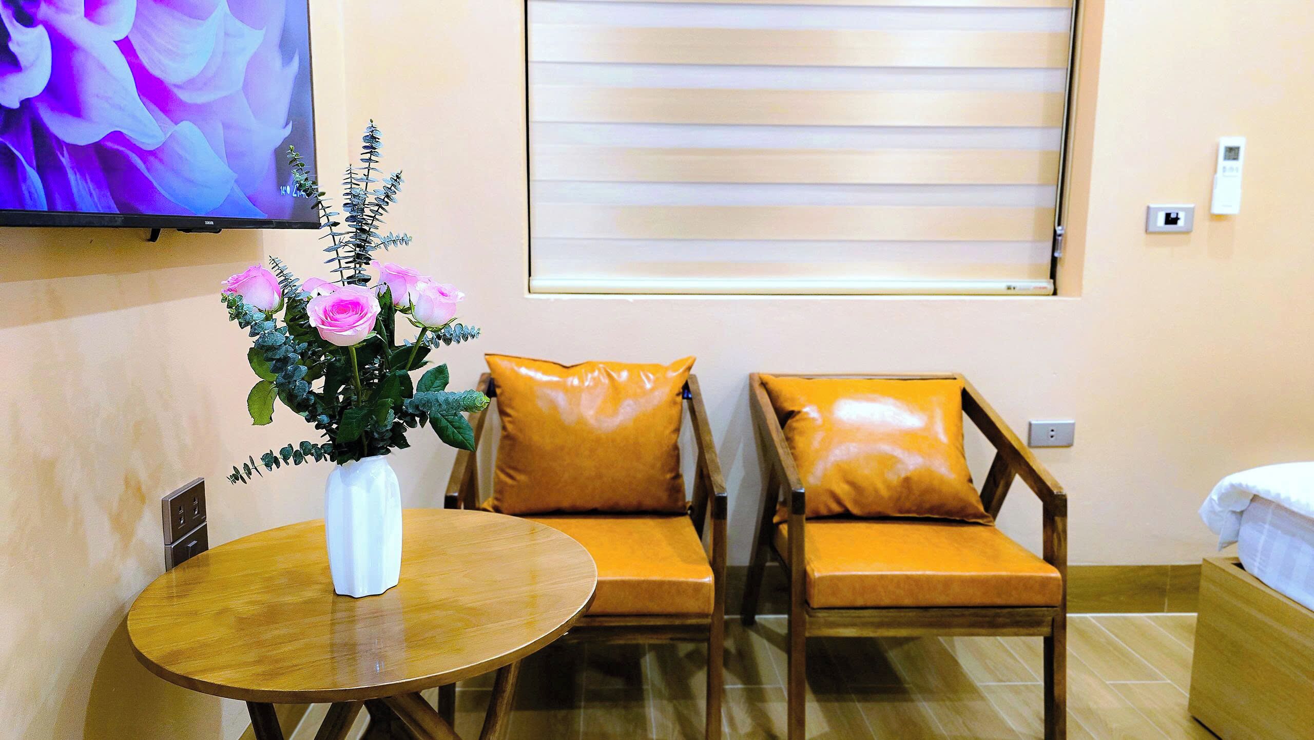 Habitación de hotel con una mesa de madera, dos sillones de cuero, un florero con flores rosas, un televisor en la pared y una ventana con persianas.
