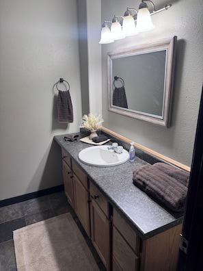 A bathroom with a white sink, dark countertop, wooden cabinets, a mirror, and a towel rack. - Hilltop Hideaway~ Seclusion in the Last Frontier (Seward)