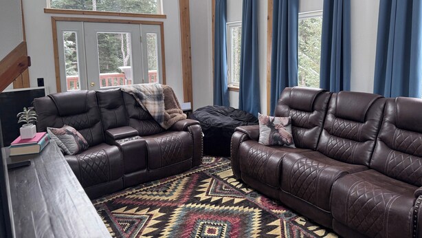 Ein gemütliches Wohnzimmer mit Ledersofas, einem gemusterten Teppich und einem großen Fenster mit Vorhängen.