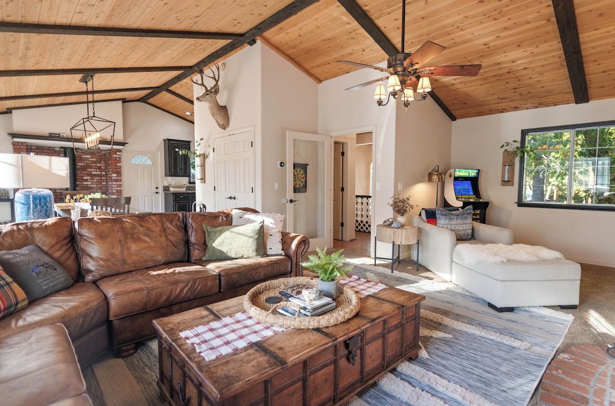A spacious living room with a brown leather sofa, a white armchair, and a wooden coffee table. A ceiling fan and a mounted deer head are visible.