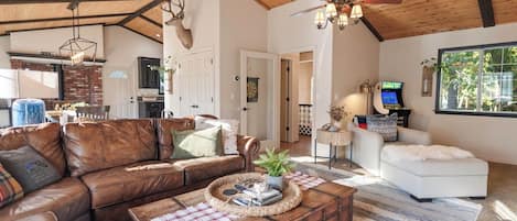 A spacious living room with a brown leather sofa, a white armchair, and a wooden coffee table. A ceiling fan and a mounted deer head are visible.