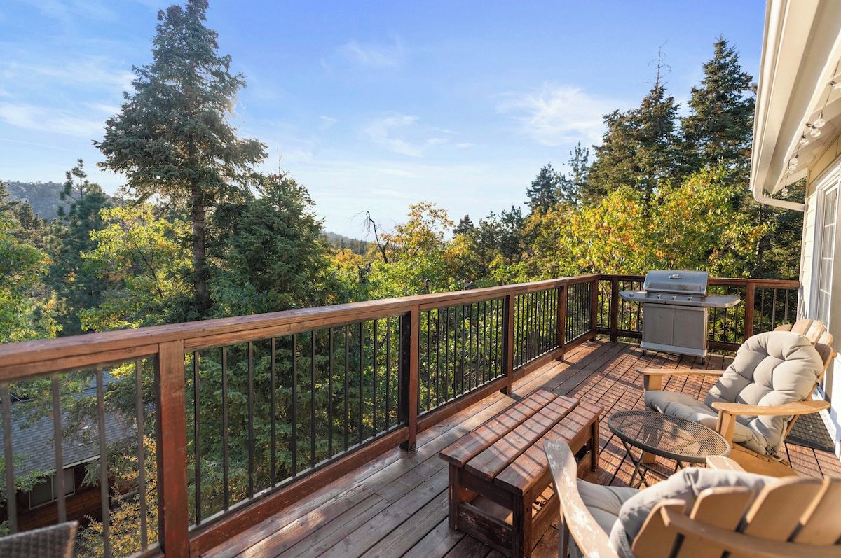 Una terraza de madera con mobiliario exterior, una parrilla y vistas a un área boscosa.