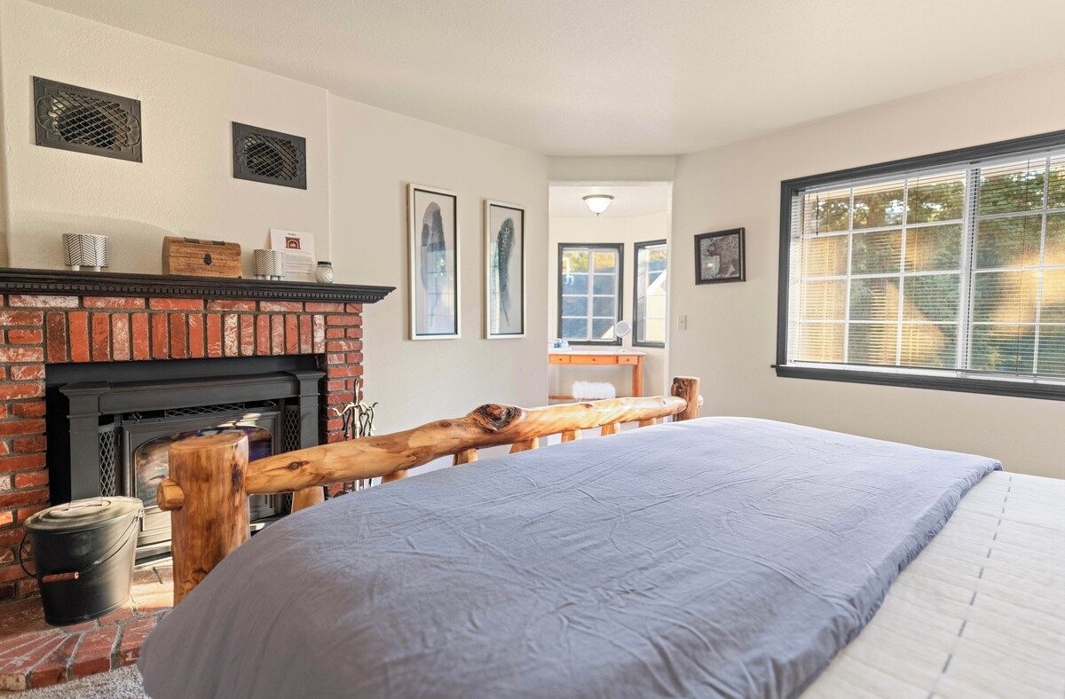 A bedroom with a brick fireplace, a bed with a blue bedspread, a wooden bench, and a large window with blinds.