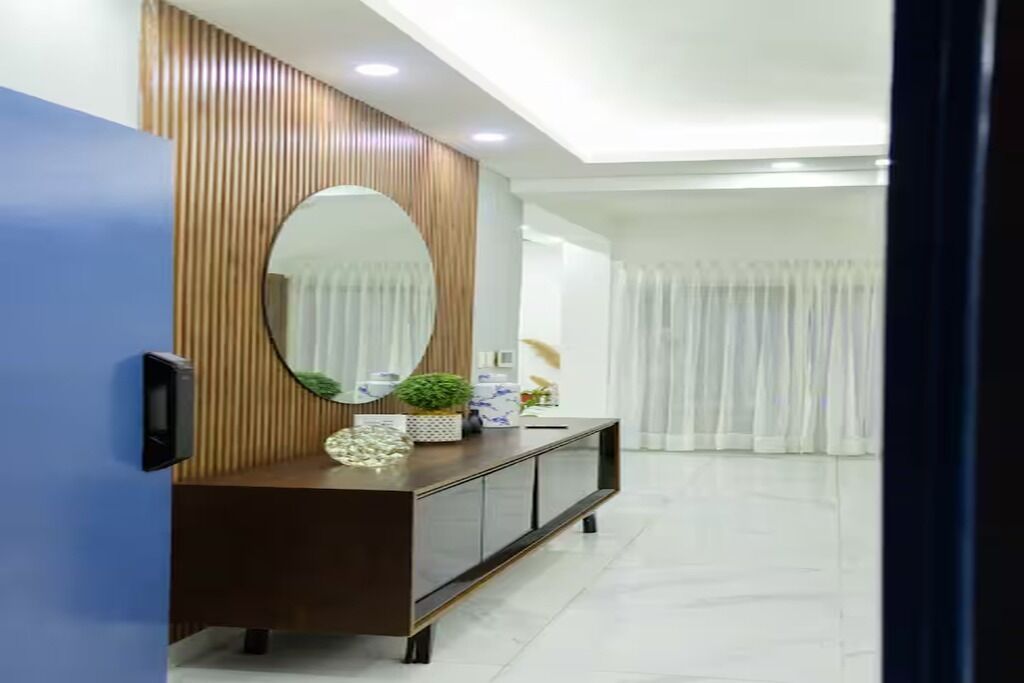 A modern living room with a wooden panel wall, a large oval mirror, a dark brown console table with decorative items, and a white curtain in the background.