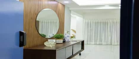 A modern living room with a wooden panel wall, a large oval mirror, a dark brown console table with decorative items, and a white curtain in the background.