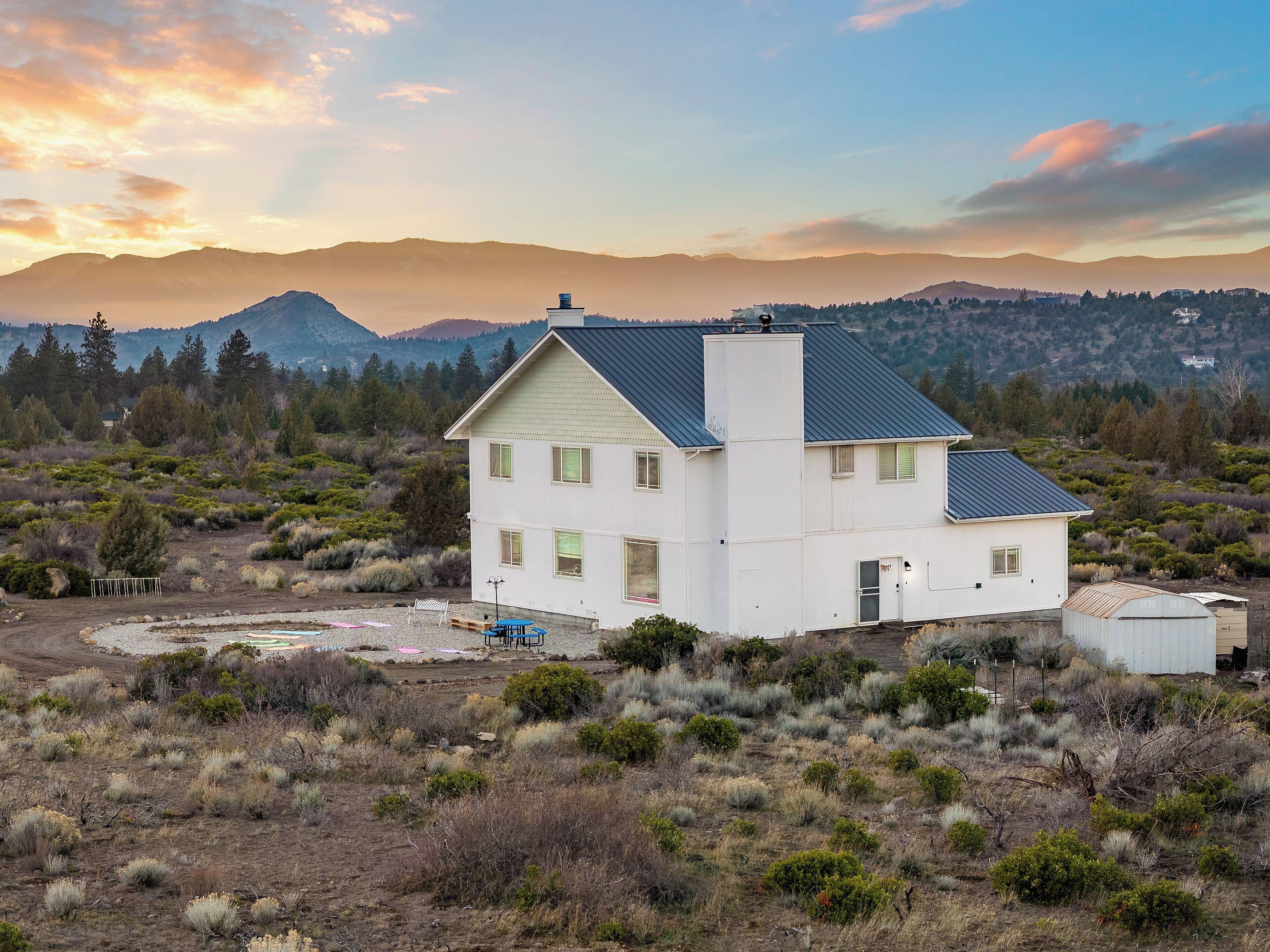 Une maison blanche à deux étages, surmontée d’un toit sombre, entourée de broussailles sèches et avec une chaîne de montagnes en arrière-plan.
