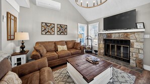 Ein Wohnzimmer mit braunem Ledersofa, einem Couchtisch, einem Kamin mit Steinverkleidung, einem flachbildfernseher über dem Kamin und einer Kristallleuchtre.