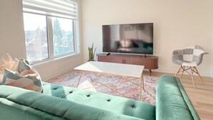 Moderni olohuone, jossa on suuri ikkuna, litteänäyttöinen televisio, vihreä sohva ja pieni pöytä.