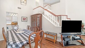 Un comedor con mantel a cuadros, sillas de madera y un armario. Una escalera con barandillas blancas conduce a otro nivel.