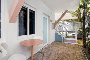 Une petite terrasse extérieure avec une table ronde et deux chaises, une porte blanche et un escalier menant à un niveau supérieur.