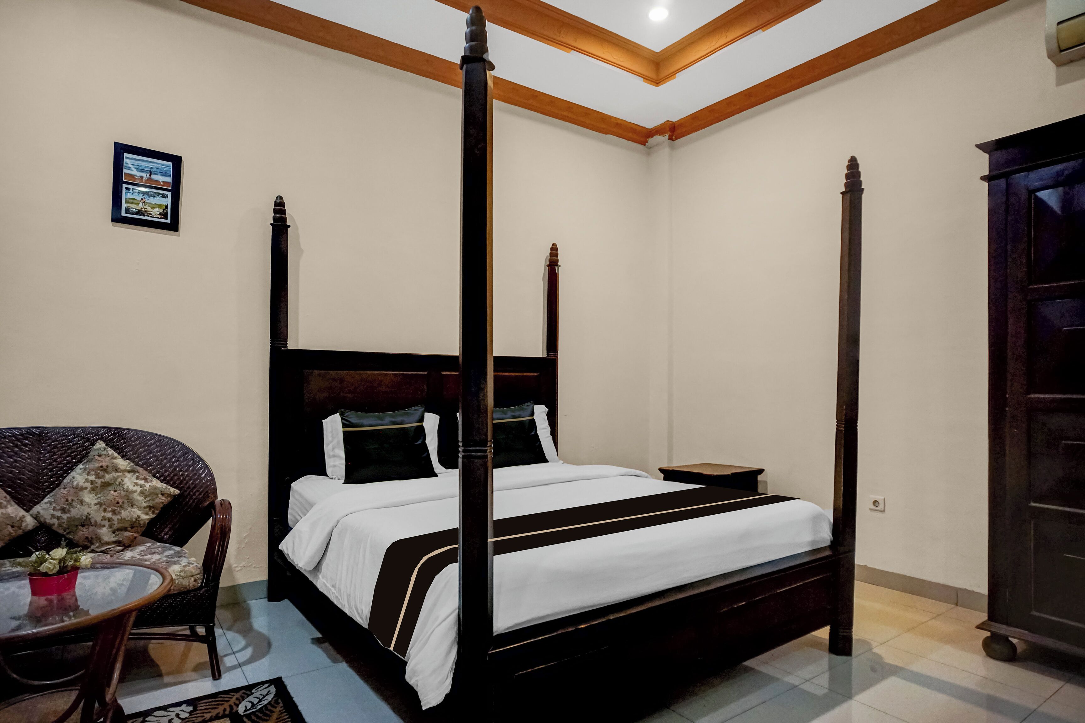 A four-poster bed with a dark wooden frame, a single sofa with floral cushions, a small round table with a red pot, and a dark wooden wardrobe.