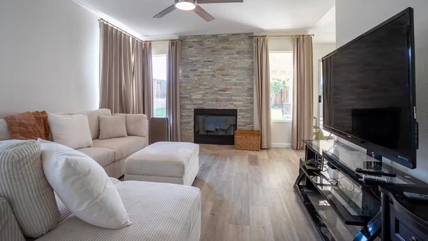 A modern living room with a stone fireplace, a large flat-screen TV, and a ceiling fan. - Spacious 4-bedroom house in superb West Palmdale. Close to the mall. (Palmdale)