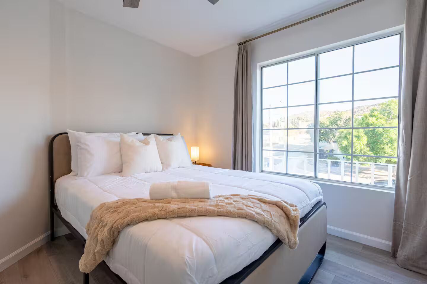 A neatly made bed with white linens and a beige throw blanket, a bedside lamp, and a large window with a view of greenery.