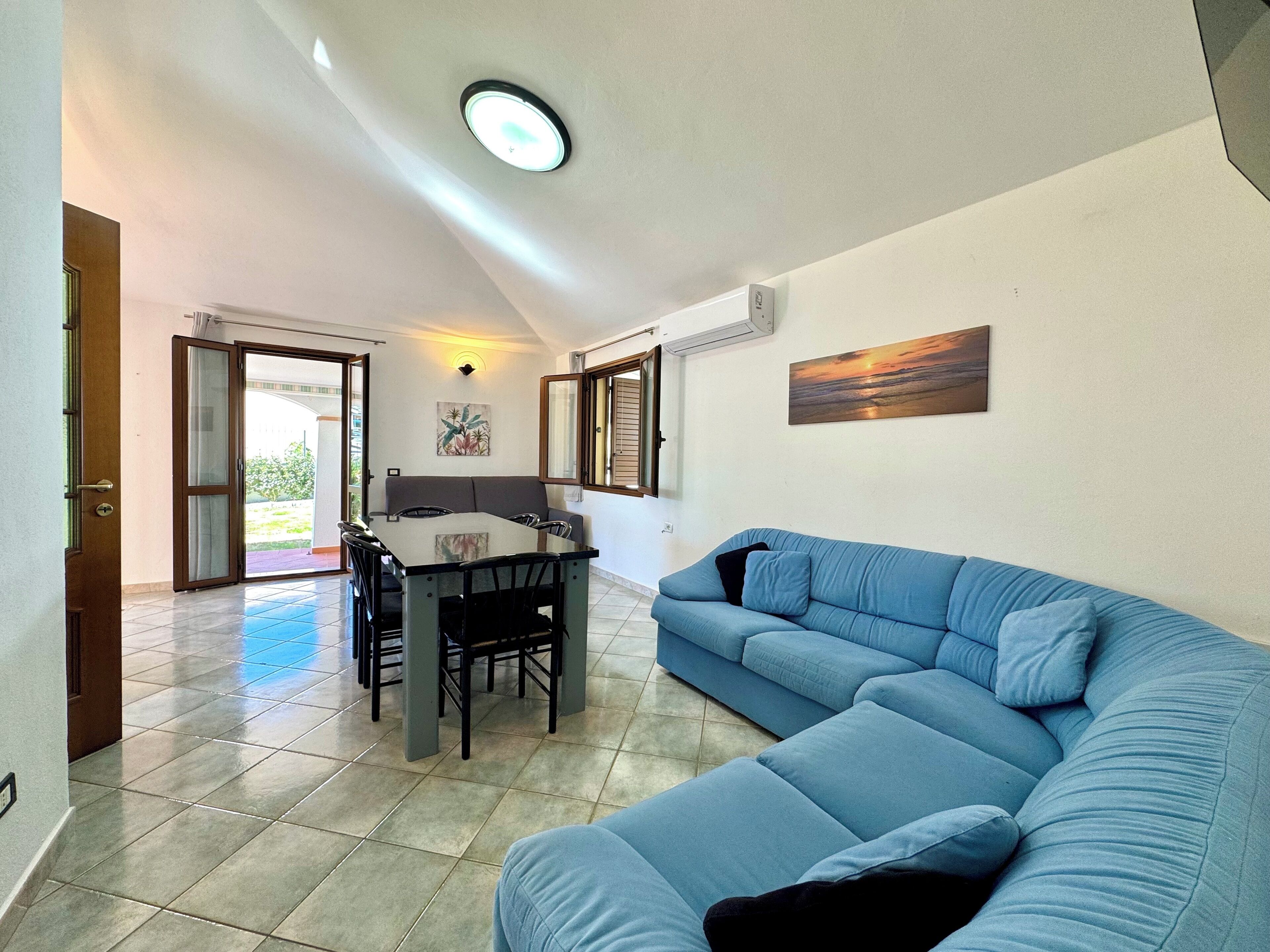Uma sala de estar com um sofá azul, uma mesa de jantar com cadeiras e uma luminária no teto.