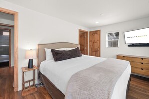 A bedroom with a bed, a bedside table with a lamp, a TV mounted on the wall, and wooden doors.