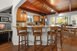 A kitchen with wooden cabinets, a bar area with stools, and a dining table. - Mountain Retreat–Steps To Bear Mountain Ski (Big Bear Lake)