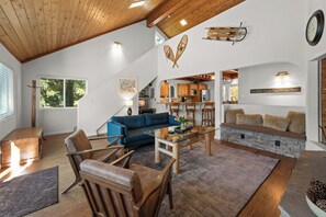 A spacious living area with a blue sofa, wooden chairs, a glass coffee table, and a stone fireplace. - Mountain Retreat–Steps To Bear Mountain Ski (Big Bear Lake)