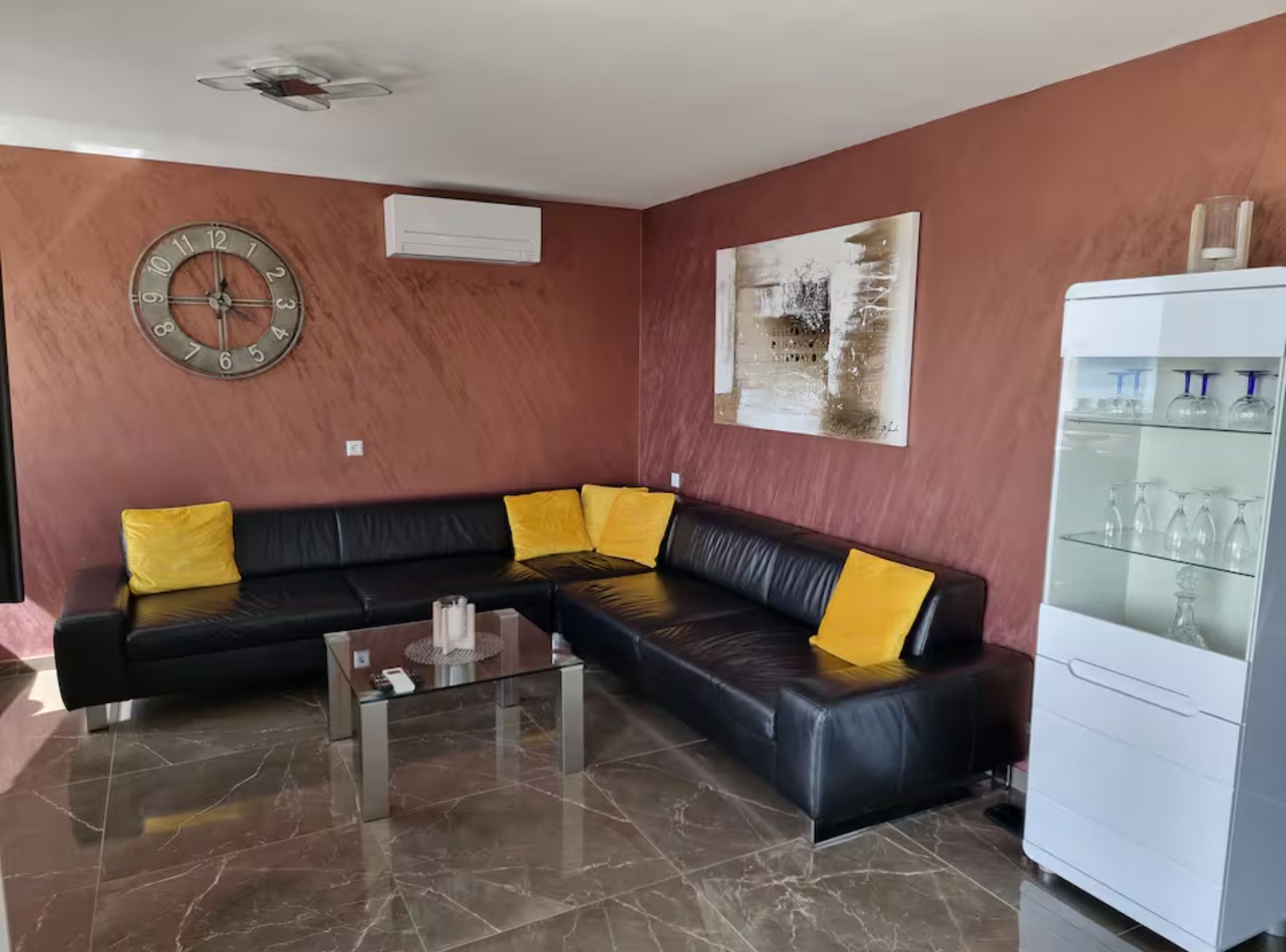 A living room with a black leather sofa, yellow cushions, a glass coffee table, a wall clock, an air conditioning unit, a painting, and a white cabinet with glassware.