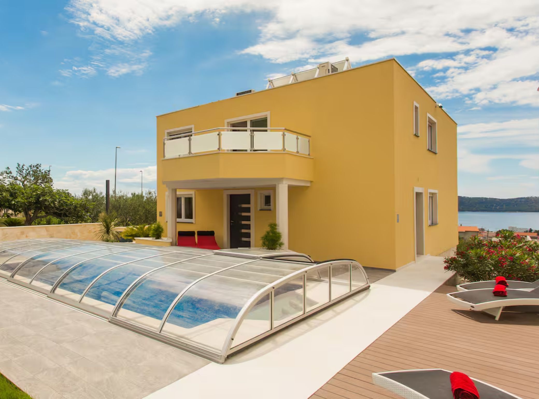 Un edificio moderno de dos pisos con techo plano, piscina cubierta y terraza de madera con sillas de descanso.