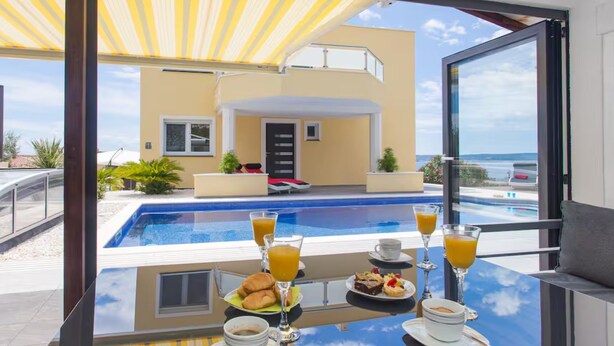 A balcony with a pool view, breakfast setup, and a modern building in the background. - Villa Rocco del Mare (Kastela)