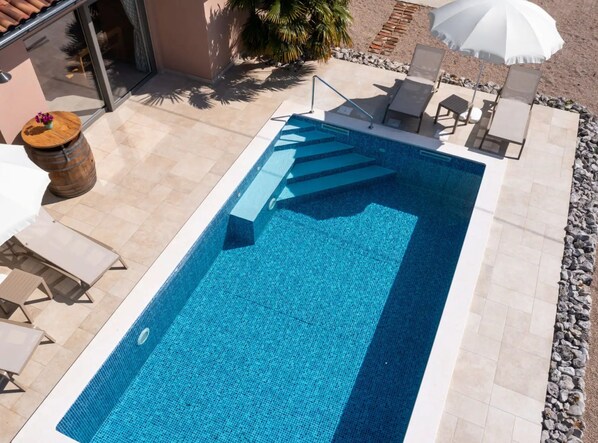 A rectangular swimming pool with a clear blue tile interior, surrounded by a stone deck. There are lounge chairs and a white umbrella providing shade. - Villa Ulika With Pool (Malinska-Dubasnica)