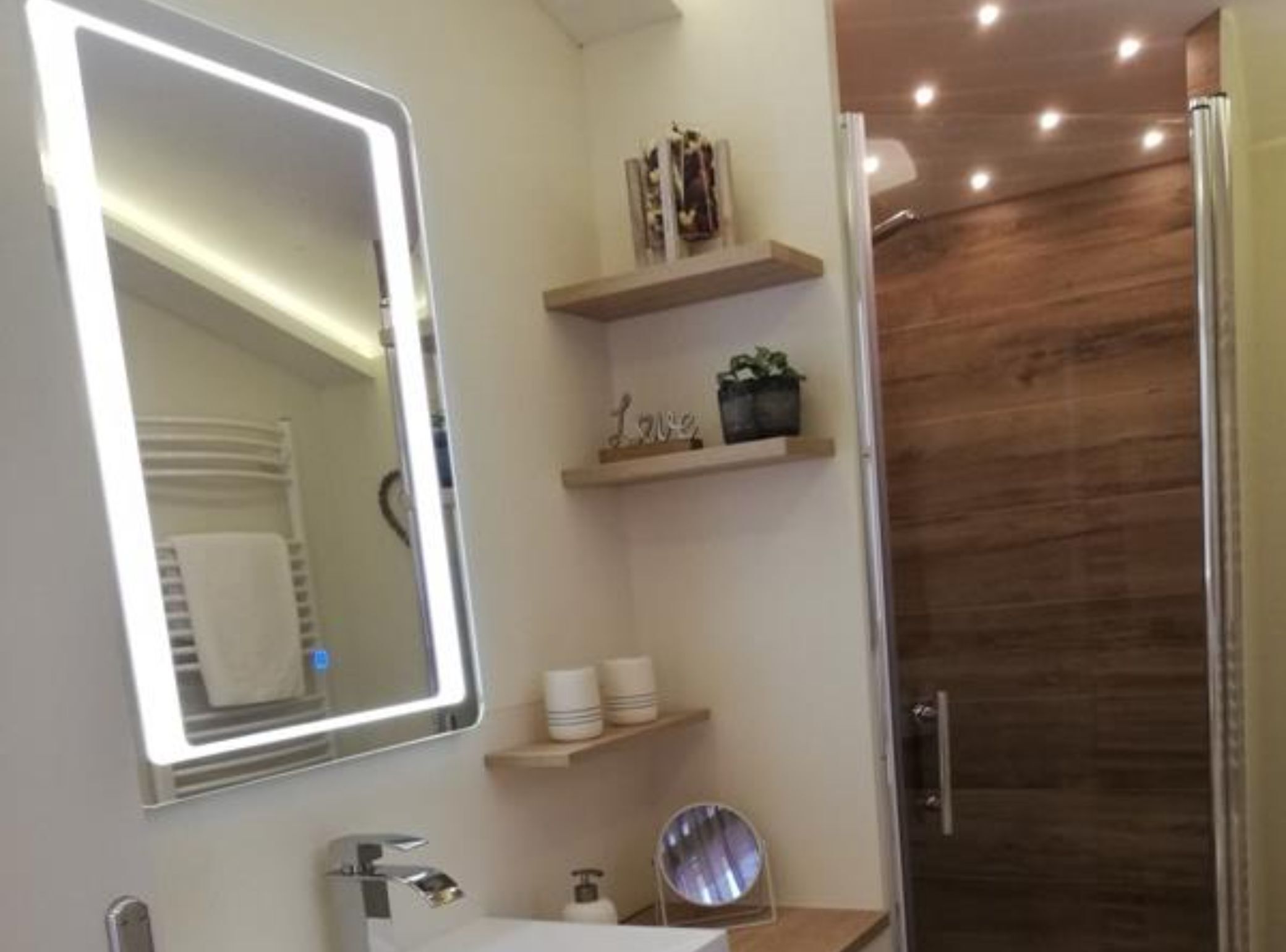 A modern bathroom with a large mirror, a shower, and wooden shelving with toiletries.