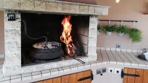 A stone-built outdoor fireplace with a fire burning inside, surrounded by a tiled floor and a wooden bench. - Holiday Home Luka Topličica (Novi Marof)
