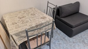 A small room with a metal-framed table covered with a tablecloth featuring food and drink illustrations, a grey sofa, and a tiled floor. - Apartments Boris-ivan (Marina)
