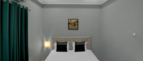 Quarto com cama, cortinas verdes, uma luminária na parede e uma luminária no teto, além de um pequeno quadro.