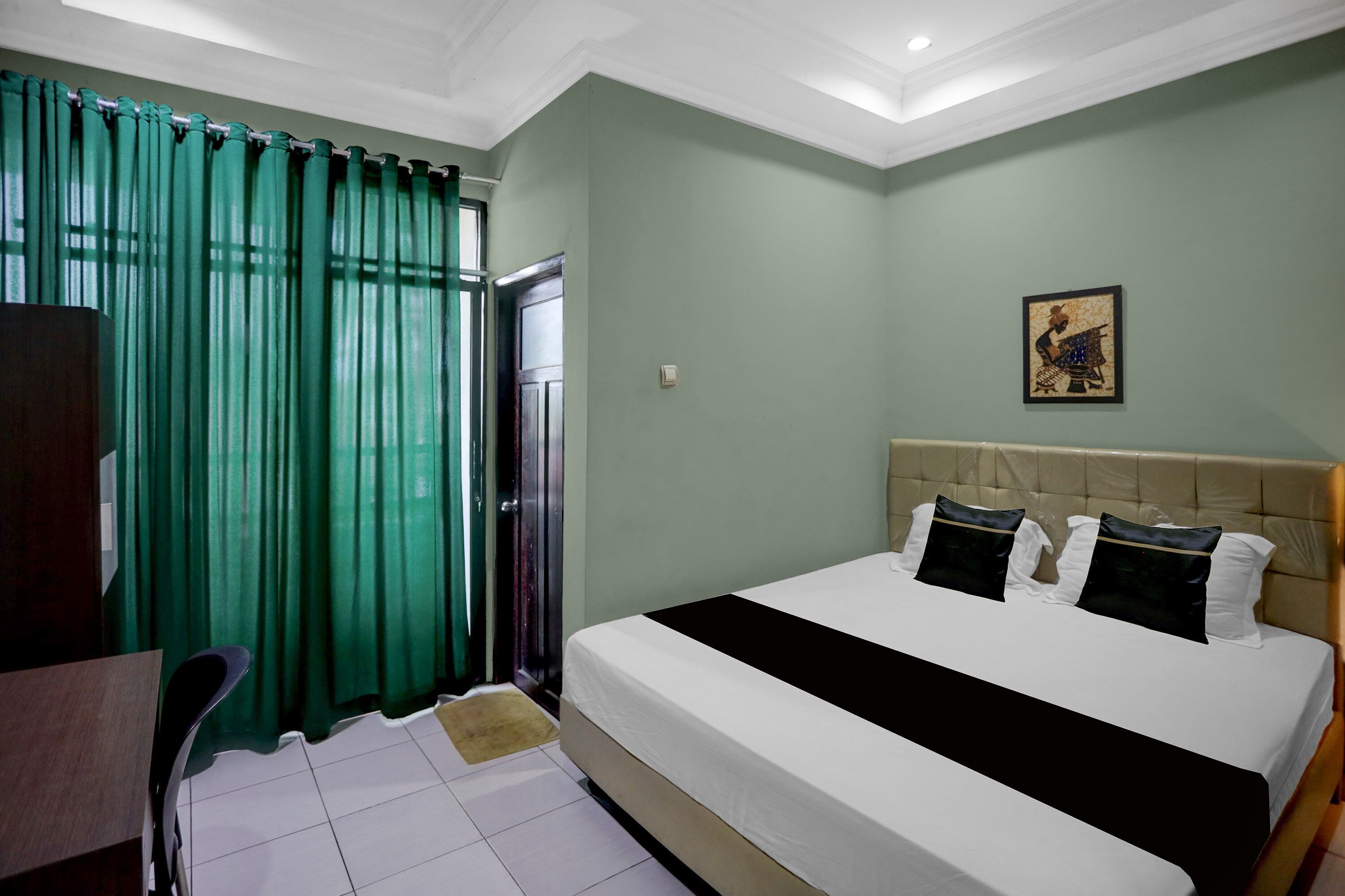 Habitación de hotel con cama, escritorio, silla, ventana con cortinas e imagen colgada en la pared.