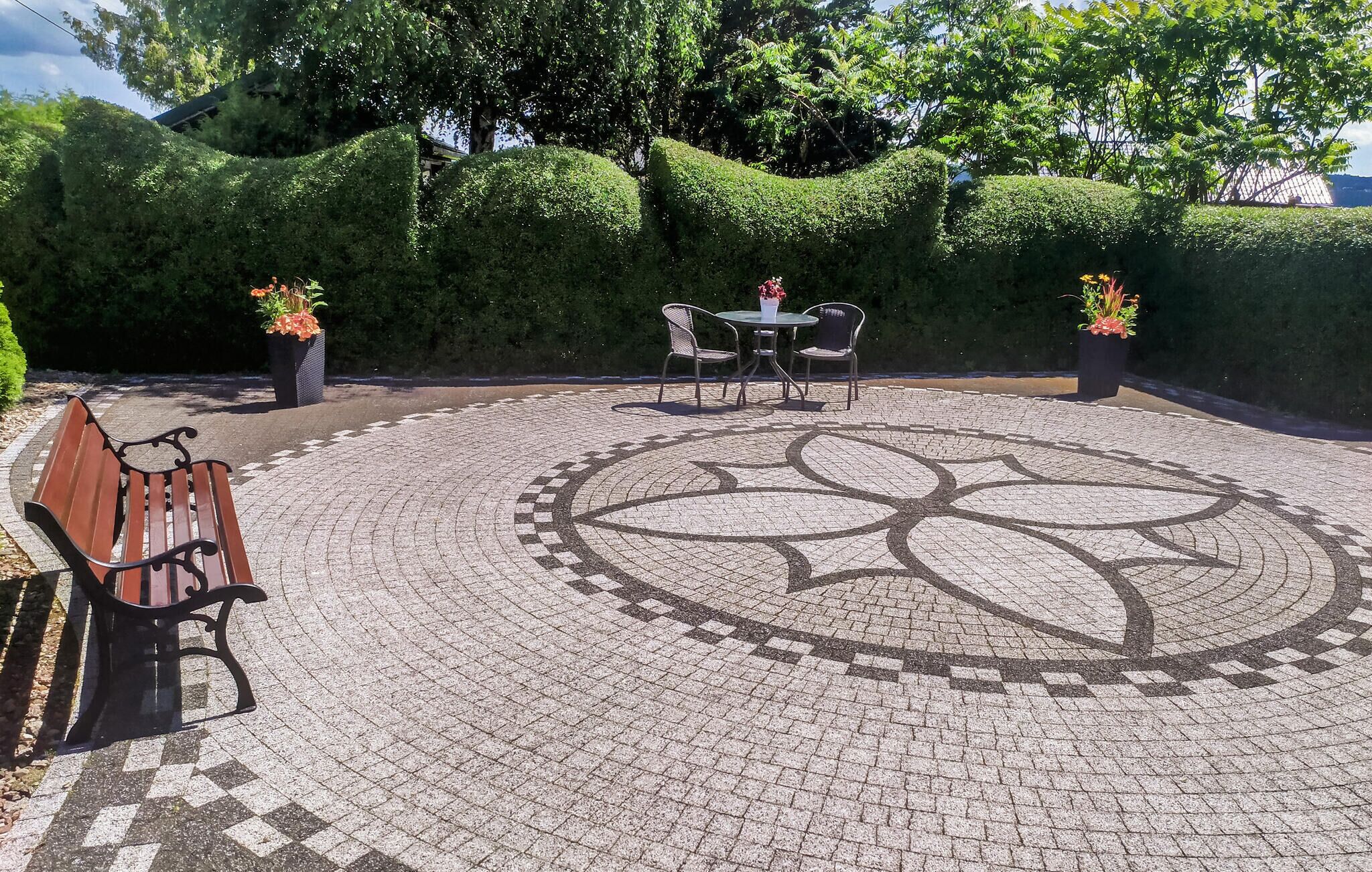 A circular outdoor seating area with a patterned floor, surrounded by neatly trimmed hedges and potted plants.