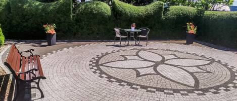 A circular outdoor seating area with a patterned floor, surrounded by neatly trimmed hedges and potted plants.