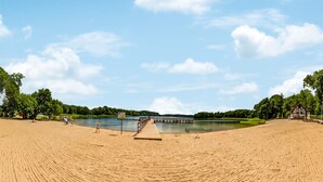Une plage avec une jetée en bois qui s’avance sur un lac paisible, entourée de verdure et sous un ciel dégagé.