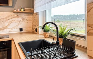 Uma cozinha com backsplash de mármore, pia preta com torneira, escorredor de louça e janela com vista para uma floresta.