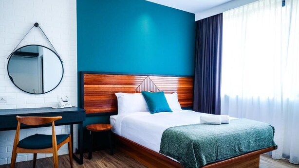 A hotel room with a wooden bed, a desk, a chair, a round mirror, and a blue accent wall. - The Journey Hotel Penang (George Town)