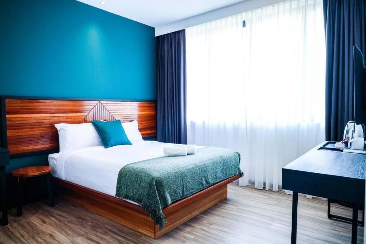 Una habitación de hotel moderna con cabecera de madera, cama bien hecha con ropa de cama blanca y almohadas color turquesa, una mesita de noche pequeña con lámpara y vistas de la ciudad a través de cortinas de gasa.