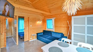 Interior acolhedor de cabana com um sofá azul, uma mesa de jantar com tampo branco e almofadas cinzas, e uma televisão montada na parede.